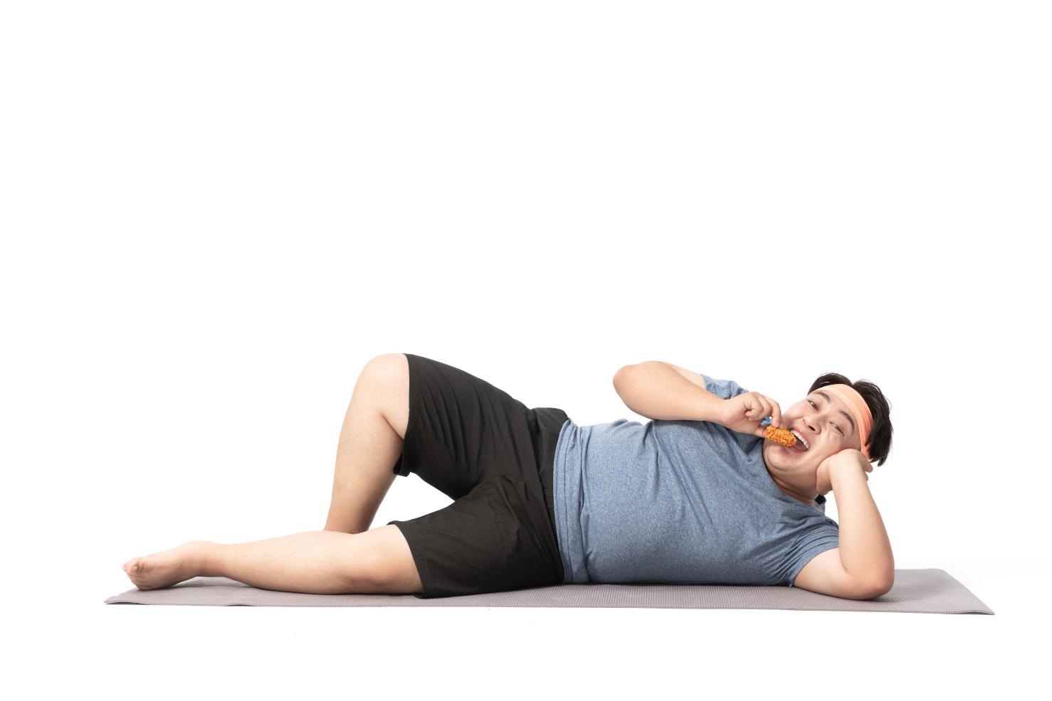 摄图网_501658088_肥胖男生躺在瑜伽垫上吃零食（非企业商用）